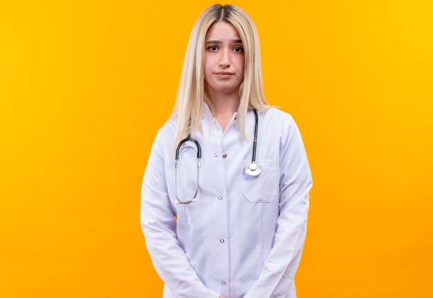 junges Mädchen des Doktors, das Stethoskop im medizinischen Kleid auf isolierter gelber Wand trägt