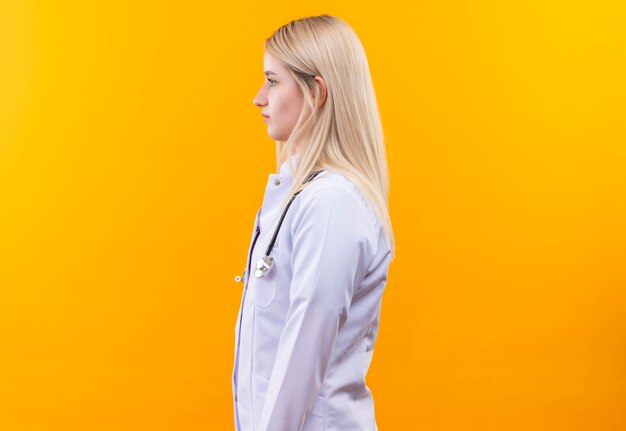 Junges Mädchen des Doktors, das Stethoskop im medizinischen Kleid trägt, das Seite auf isolierte gelbe Wand betrachtet