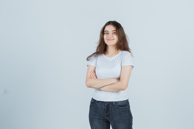 Junges Mädchen hält Arme gefaltet in T-Shirt, Jeans und schaut erfreut. Vorderansicht.