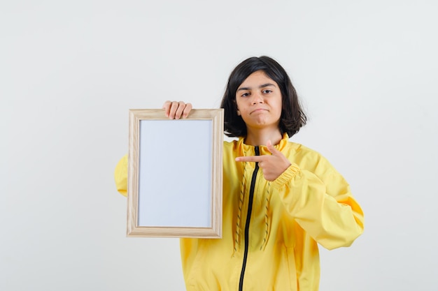 Junges Mädchen hält Whiteboard, zeigt darauf mit Zeigefinger in gelber Bomberjacke und sieht ernst aus.