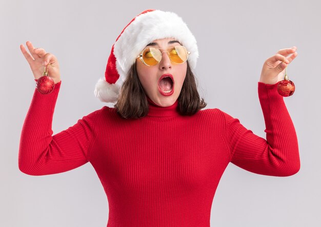 Junges Mädchen im roten Pullover und in der Weihnachtsmannmütze, die die Brille hält, die Weihnachtskugeln hält, die beiseite überrascht über weißem Hintergrund stehen
