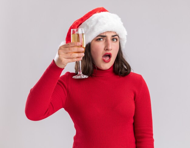 Junges Mädchen im roten Pullover und in der Weihnachtsmannmütze, die Glas Champagner hält, überrascht, über weißer Wand stehend