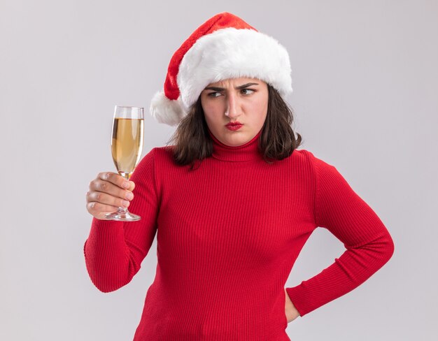 Junges Mädchen im roten Pullover und in der Weihnachtsmannmütze, die Glas Champagner halten, das betrachtet, dass es unzufrieden ist, über weißer Wand zu stehen