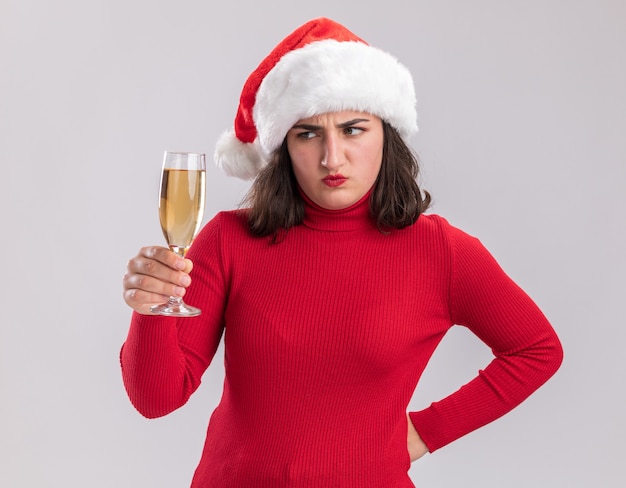 Junges Mädchen im roten Pullover und in der Weihnachtsmannmütze, die Glas Champagner halten, das betrachtet, dass es unzufrieden ist, über weißer Wand zu stehen