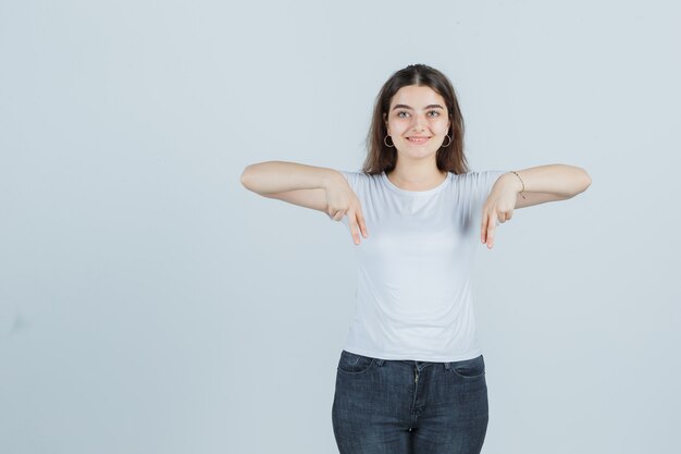 Junges Mädchen im T-Shirt, Jeans, die nach unten zeigen und glücklich schauen, Vorderansicht.