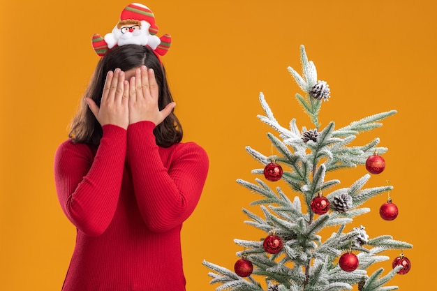 Junges Mädchen im Weihnachtspullover, der lustiges Stirnband trägt, das Augen mit den Händen steht, die neben einem Weihnachtsbaum über orange Hintergrund stehen