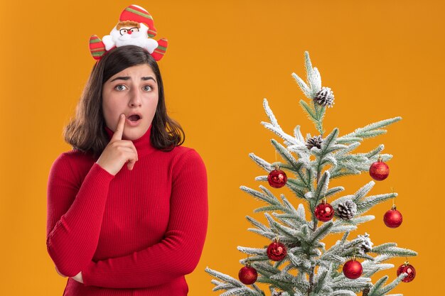 Junges Mädchen im Weihnachtspullover, der lustiges Stirnband trägt, das die Kamera überrascht, die neben einem Weihnachtsbaum über orange Hintergrund steht