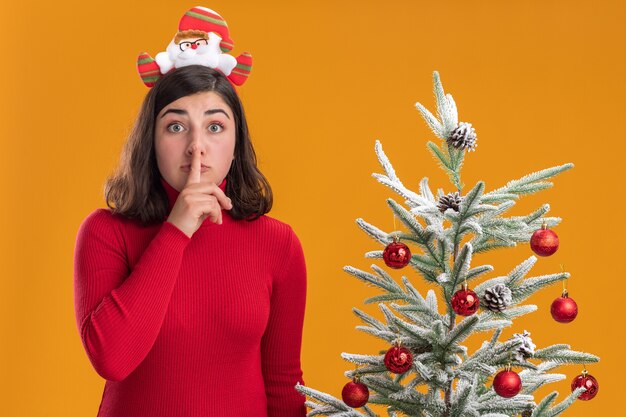 Junges Mädchen im Weihnachtspullover, der lustiges Stirnband trägt, das Kamera betrachtet, die Stille Geste nahe einem Weihnachtsbaum über orange Hintergrund macht