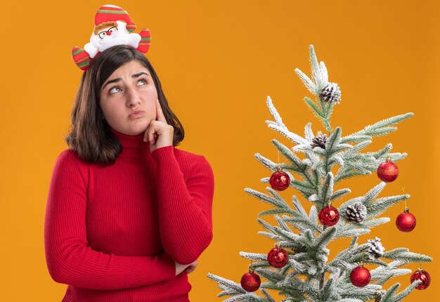 Junges Mädchen im Weihnachtspullover, der lustiges Stirnband trägt, das verwirrt neben einem Weihnachtsbaum über orange Hintergrund schaut