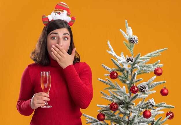 Junges Mädchen im Weihnachtspullover, der lustiges Stirnband trägt und Glas Champagner neben einem Weihnachtsbaum über orange Hintergrund hält