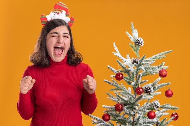 Junges Mädchen im Weihnachtspullover, der lustiges und aufgeregtes lustiges Stirnband neben einem Weihnachtsbaum über orange Hintergrund trägt