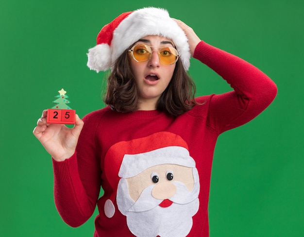 Junges Mädchen im Weihnachtspullover, der Weihnachtsmütze und Brille hält, die Spielzeugwürfel mit Nummer fünfundzwanzig überrascht und erstaunt über grüner Wand stehen