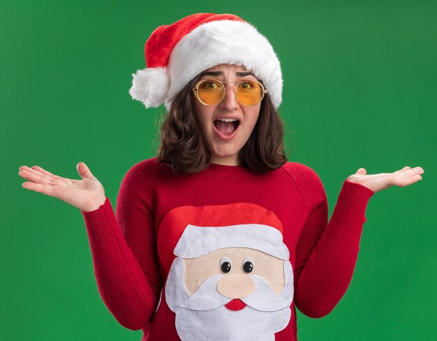 Junges Mädchen im Weihnachtspullover, der Weihnachtsmütze und Brille trägt verwirrt Hände, die über grüner Wand stehen