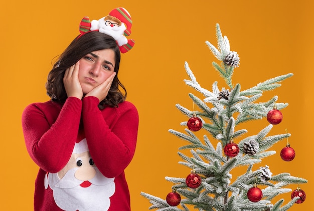 Junges Mädchen im Weihnachtspullover mit lustigem Stirnband, das mit traurigem Gesichtsausdruck neben einem Weihnachtsbaum auf orangefarbenem Hintergrund in die Kamera schaut