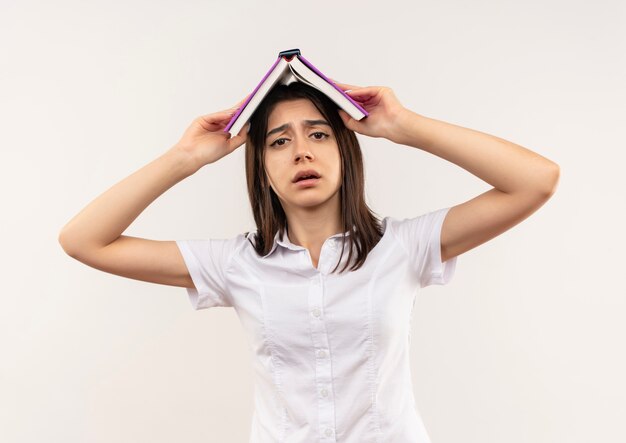 Junges Mädchen im weißen Hemd, das offenes Buch über ihrem Kopf hält und verwirrt steht über weißer Wand