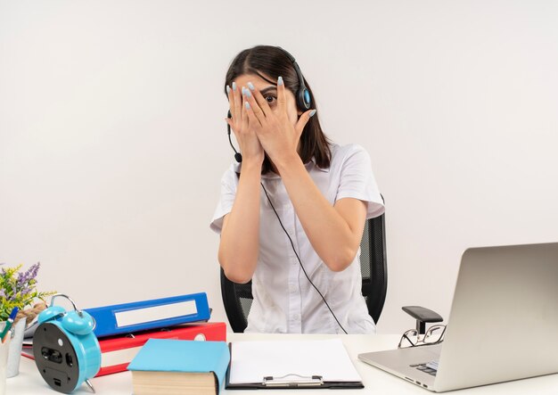 Junges Mädchen im weißen Hemd und in den Kopfhörern, verängstigte bedeckende Augen mit Handflächen, die durch Finger schauen, die am Tisch mit Ordnern und Laptop über weißer Wand sitzen