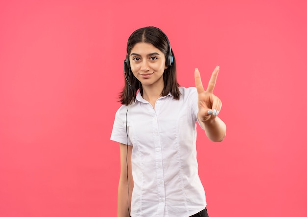 Junges Mädchen im weißen Hemd und in den Kopfhörern, zeigend und zeigend mit den Fingern oben Nummer zwei lächelnd stehend über rosa Wand