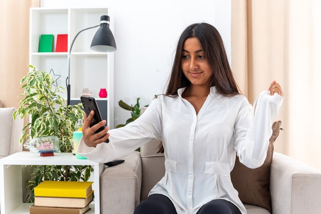 Junges Mädchen im weißen Hemd und in der schwarzen Hose, die die geballte Faust des Smartphones halten glücklich und fröhlich sitzen auf dem Stuhl im hellen Wohnzimmer sitzen