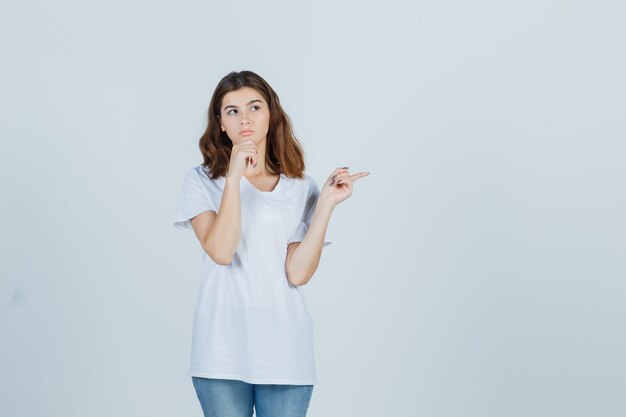 Junges Mädchen im weißen T-Shirt, das beiseite zeigt, Kinn auf Hand stützt und nachdenklich, Vorderansicht schaut.