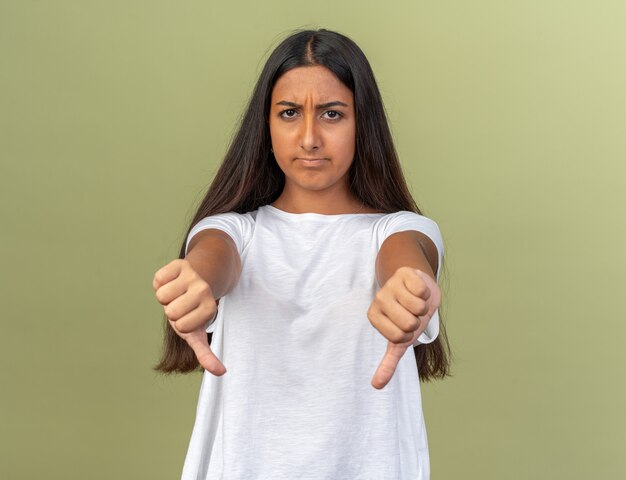 Junges Mädchen im weißen T-Shirt, das die Kamera anschaut, ist unzufrieden und zeigt Daumen nach unten, die über Grün stehen