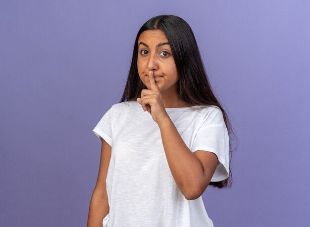 Junges Mädchen im weißen T-Shirt, das die Kamera anschaut und eine Stille-Geste mit dem Finger auf den Lippen macht, die auf blauem Hintergrund stehen