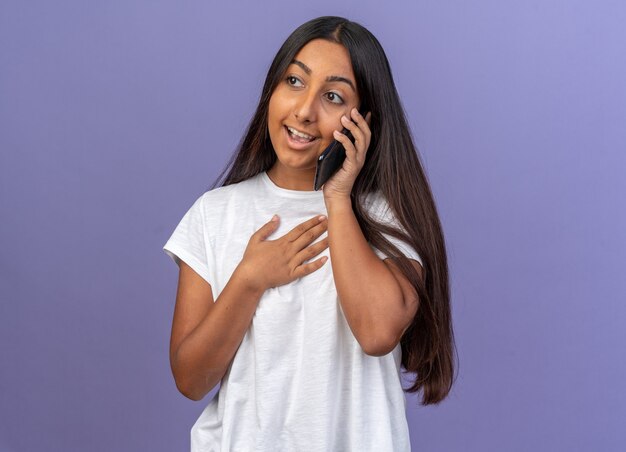 Junges Mädchen im weißen T-Shirt, das fröhlich lächelt, während es auf dem Handy spricht, das auf blauem Hintergrund steht