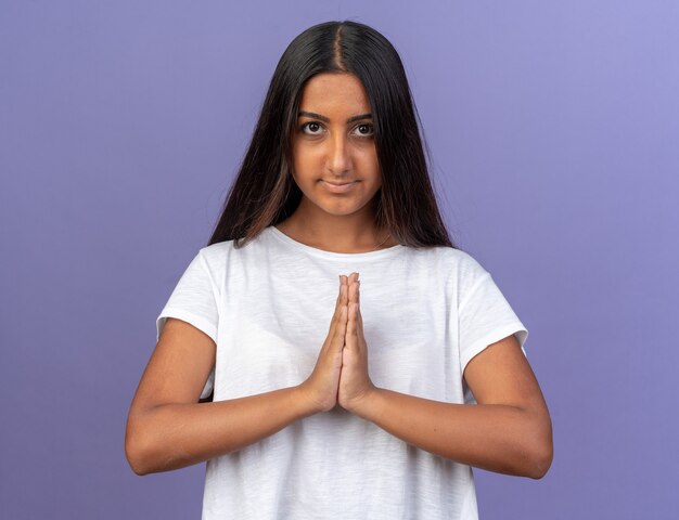 Junges Mädchen im weißen T-Shirt, das Händchen hält wie eine Namaste-Geste, die mit einem Lächeln im Gesicht in die Kamera schaut