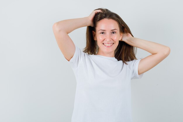 Junges Mädchen im weißen T-Shirt, das mit den Händen im Haar aufwirft und charmant, Vorderansicht schaut.