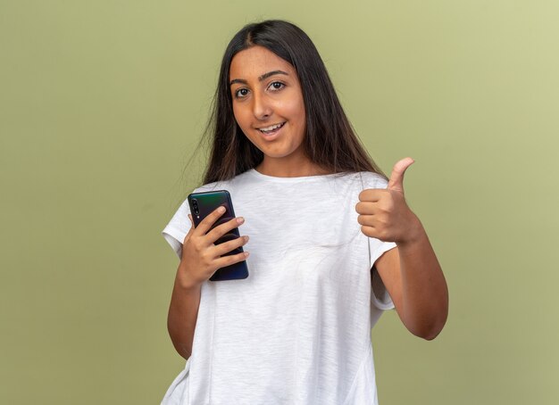 Junges Mädchen im weißen T-Shirt, das Smartphone mit Blick in die Kamera hält und mit glücklichem Gesicht lächelt