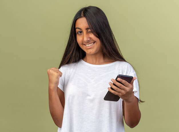 Junges Mädchen im weißen T-Shirt, das Smartphone mit geballter Faust glücklich und aufgeregt lächelnd und zwinkernd über grünem Hintergrund hält