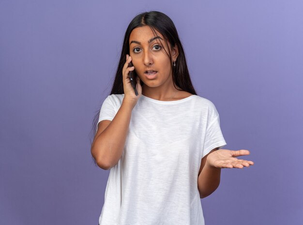 Junges Mädchen im weißen T-Shirt, das verwirrt aussieht, während es auf dem Handy spricht, das auf blauem Hintergrund steht