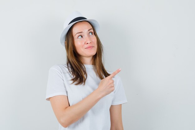 Junges Mädchen im weißen T-Shirt, Hut zeigt auf obere rechte Ecke und schaut fokussiert, Vorderansicht.