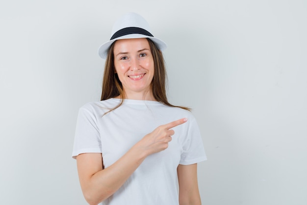 Junges Mädchen im weißen T-Shirt, Hut zeigt auf obere rechte Ecke und schaut froh, Vorderansicht.