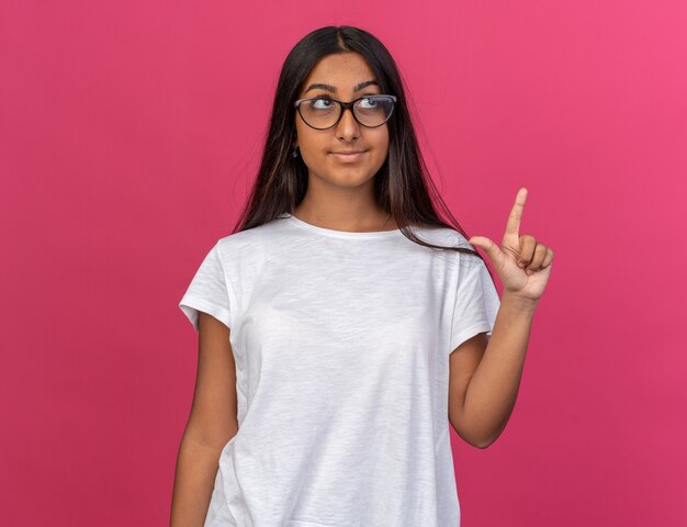 Junges Mädchen im weißen T-Shirt mit Brille, das mit einem Lächeln auf dem Gesicht aufschaut und den Zeigefinger zeigt, der eine neue Idee hat, die über Pink steht?