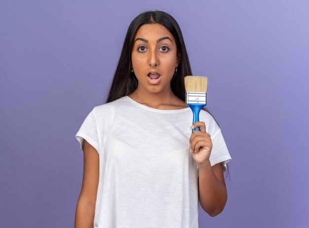 Junges Mädchen im weißen T-Shirt mit Pinsel in die Kamera schaut erstaunt und überrascht über Blau stehend