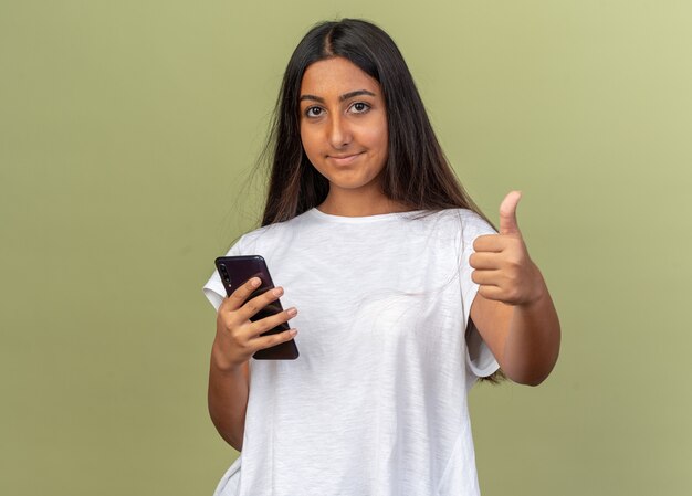 Junges Mädchen im weißen T-Shirt mit Smartphone, das lächelnd in die Kamera schaut und Daumen hoch steht über grünem Hintergrund