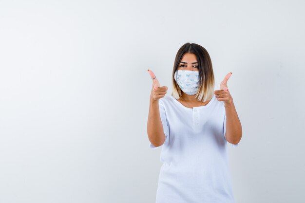 Junges Mädchen im weißen T-Shirt und in der Maske, die doppelte Daumen oben zeigt und fröhlich schaut, Vorderansicht.