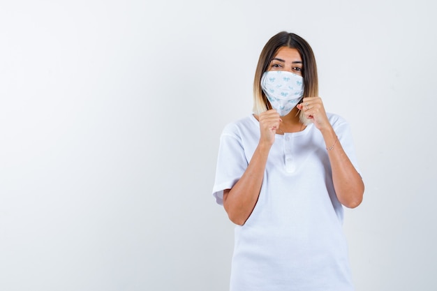 Junges Mädchen im weißen T-Shirt und in der Maske, die in der Kampfhaltung stehen und zuversichtlich schauen, Vorderansicht.