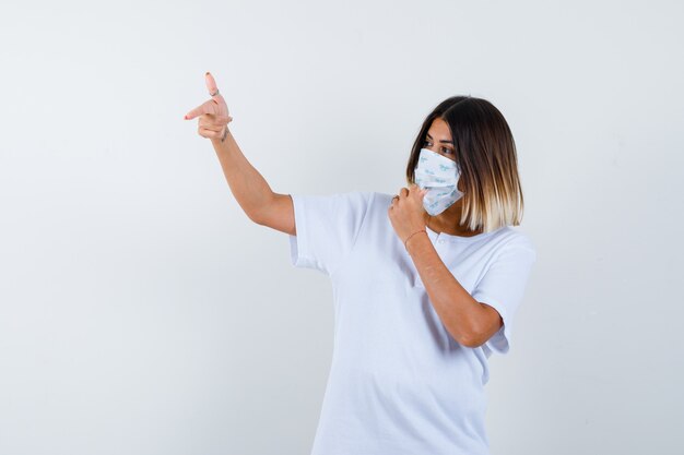 Junges Mädchen im weißen T-Shirt und in der Maske, die links mit Zeigefinger zeigen, Hand auf Mund halten und zuversichtlich, Vorderansicht schauen.