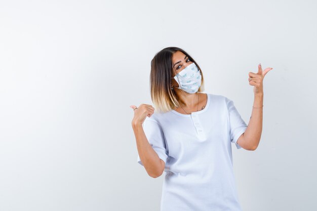 Junges Mädchen im weißen T-Shirt und in der Maske zeigt rechts und links mit den Zeigefingern und schaut fröhlich, Vorderansicht.