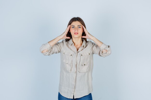 Junges Mädchen in beigefarbenem Hemd, Jeans mit Händen an den Schläfen und aufgeregt, Vorderansicht.