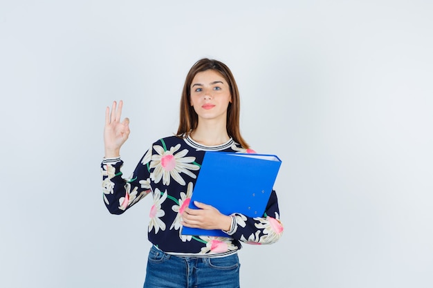 Junges Mädchen in Blumenbluse, Jeans mit Ordner, zeigt eine ok Geste und sieht selbstbewusst aus, Vorderansicht.