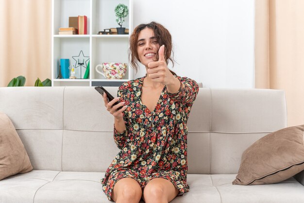 Junges Mädchen in Blumenkleid mit Smartphone lächelnd fröhlich mit Daumen nach oben auf einer Couch im hellen Wohnzimmer sitzend