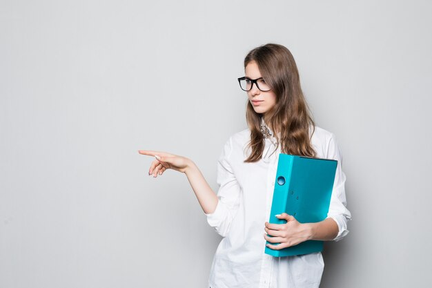 Junges Mädchen in den Gläsern, die im strengen Büro weißes T-Shirt gekleidet sind, steht vor weißer Wand mit blauem Ordner für Dokumente in ihren Händen