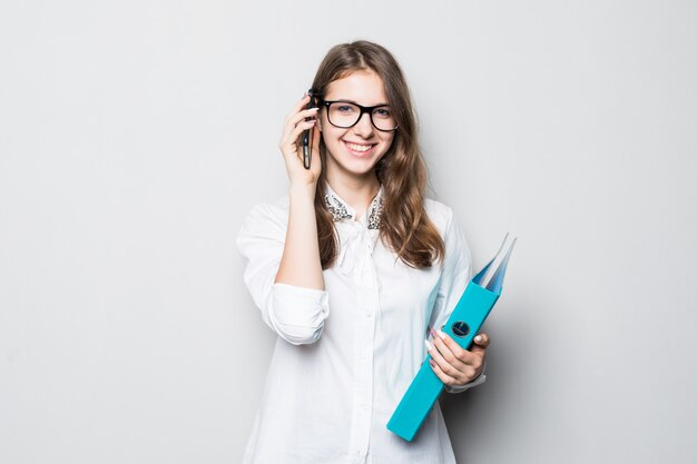 Junges Mädchen in der Brille, die im strengen Büro weißes T-Shirt gekleidet ist, steht vor weißer Wand und hält ihr Telefon und Ordner in Händen