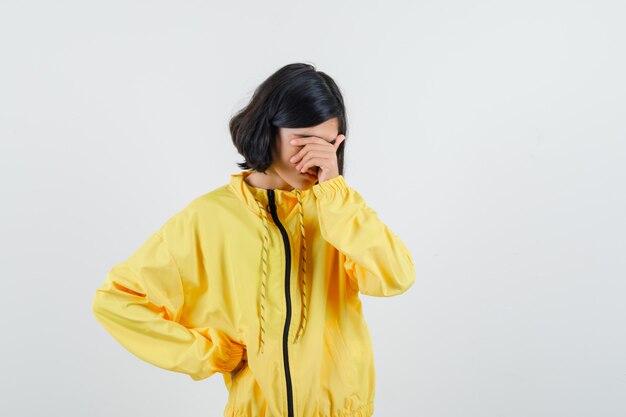 Junges Mädchen in der gelben Bomberjacke, die einen Teil des Gesichts mit der Hand bedeckt, eine andere Hand auf die Taille legt und bedauernd aussieht