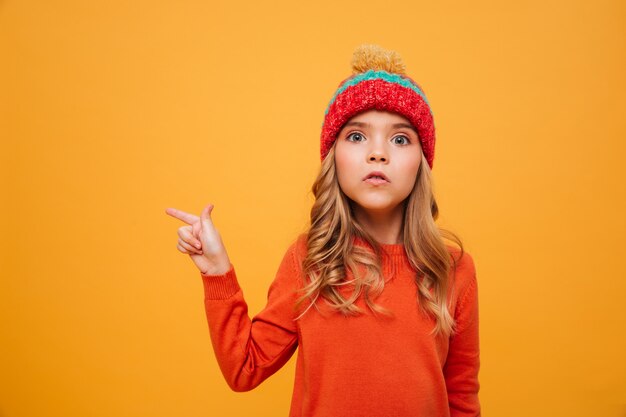 Junges Mädchen in der Strickjacke und im Hut weg zeigend und die Kamera über Orange betrachtend