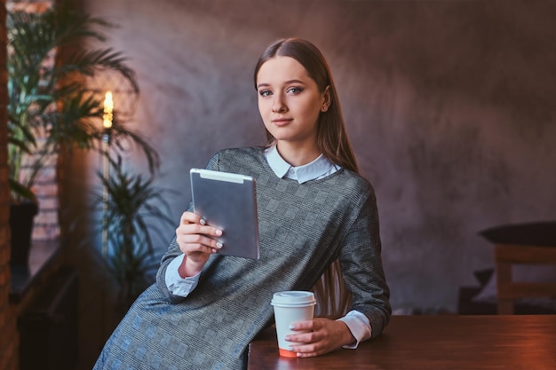 Junges Mädchen in einem eleganten grauen Kleid hält eine Tasse Kaffee zum Mitnehmen und hält ein Tablet in der Hand, während es sich in einem Raum mit Loft-Interieur auf den Tisch lehnt und in eine Kamera schaut.