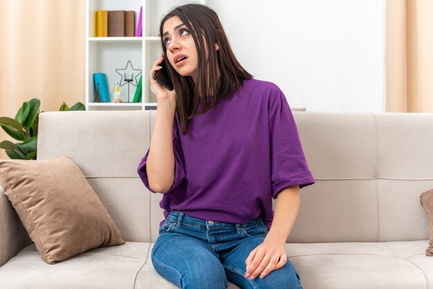 Junges Mädchen in Freizeitkleidung, das beim Telefonieren auf einer Couch im hellen Wohnzimmer genervt aussieht