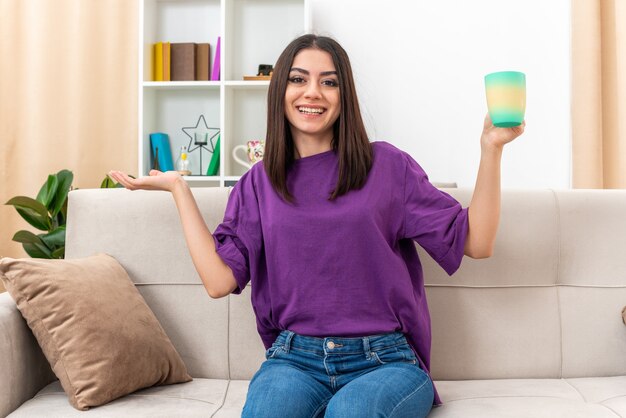 Junges Mädchen in Freizeitkleidung, das eine Tasse Tee hält, die fröhlich lächelt und etwas mit dem Arm der Hand präsentiert, das auf einer Couch im hellen Wohnzimmer sitzt